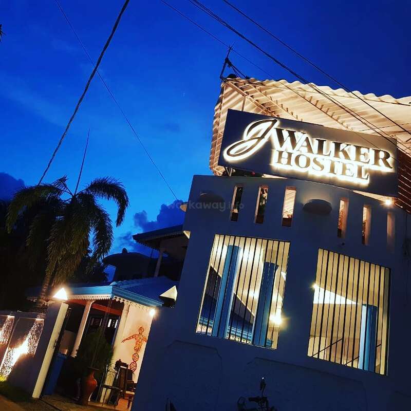 The image depicts a white building with a sign reading \"J Walker Hostel\" at dusk, featuring a palm tree and a blue sky in the background.