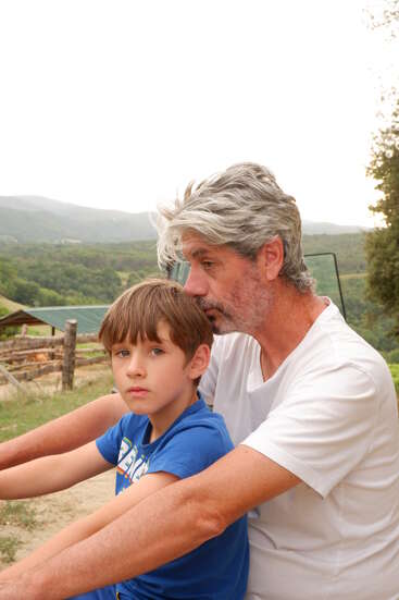 Un homme aux cheveux gris et à la barbe blanche est assis tout près derrière un jeune garçon. Tous deux semblent pensifs, entourés d'un paysage extérieur verdoyant et pittoresque.