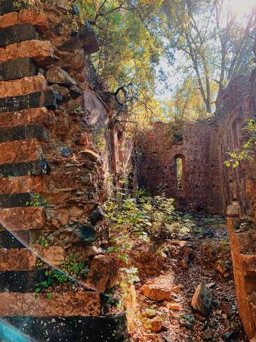 La lumière du soleil filtre à travers les arbres au-dessus des ruines de pierre anciennes. La nature reprend lentement possession de la structure en ruine, avec de la végétation qui pousse parmi les rochers tombés et une lumière chaude et tamisée qui illumine tout.