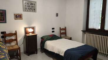 Cette chambre confortable dispose d'un lit simple, de deux chaises en bois, d'une table de nuit avec une lampe, d'œuvres d'art murales, d'une fenêtre avec des rideaux transparents et d'un décor simple.