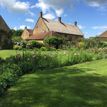 The image depicts a serene garden with vibrant flowers and lush greenery, set against the backdrop of a charming stone house with a brown roof and chimney.