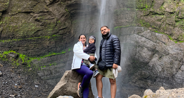 Una familia de tres miembros posa felizmente delante de una alta cascada, de pie sobre unas rocas con impresionantes acantilados al fondo, vestidos con ropa de abrigo para disfrutar de una aventura al aire libre.