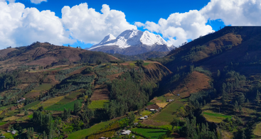 A lush green valley with scattered houses and farmland lies beneath rugged mountains. Snow-capped peaks rise majestically in the background, surrounded by fluffy white clouds.