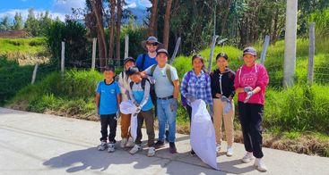 A group of children and one adult stand together outdoors, holding bags and wearing gloves, smiling, likely participating in a community clean-up event on a sunny day.