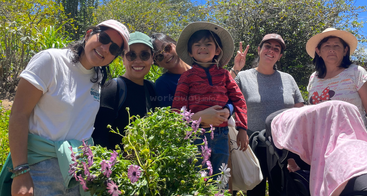 A group of smiling people, including a child, pose outdoors with flowers. They wear hats and sunglasses, enjoying a sunny day surrounded by nature and greenery.