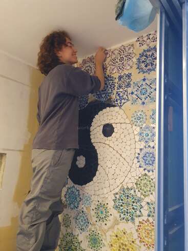 Eine Person mit lockigem Haar steht auf einer Leiter und arbeitet an einer Mosaikwand mit einem Yin-Yang-Symbol und anderen bunten Mustern auf einer weißen Wand.