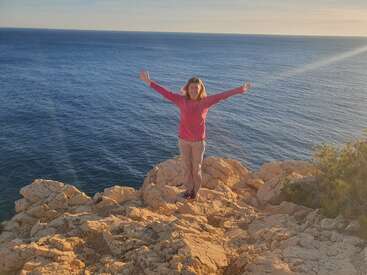 A woman stands on a rocky cliff overlooking the ocean, with her arms outstretched, wearing a pink shirt and khaki pants, smiling at the camera.