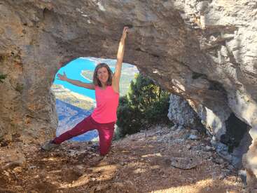A woman in bright athletic clothing poses energetically beneath a rocky archway on a mountain trail, with a turquoise lake and greenery visible in the background.