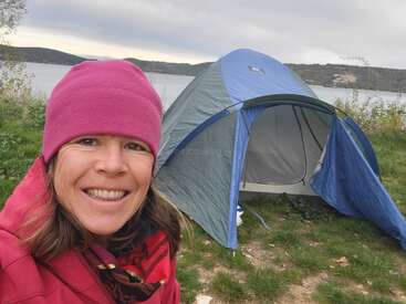 A woman in a pink hat and jacket smiles near a pitched tent beside a lake. The weather looks chilly and overcast, perfect for camping adventure.