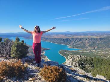 A woman stands on a rocky cliff, arms outstretched, overlooking a turquoise river and expansive mountainous landscape under a clear blue sky. Stunning natural view.
