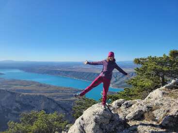 A woman in bright clothing stands joyfully on a rocky mountain ledge, overlooking a beautiful turquoise lake, surrounded by greenery under a clear blue sky.