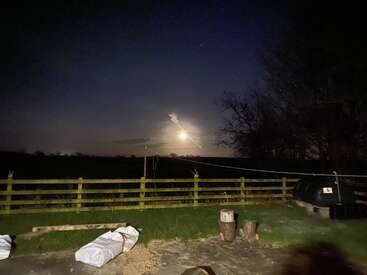 A glowing full moon lights up a dark rural landscape with a wooden fence, scattered logs, and some white covers in the grassy yard at night.