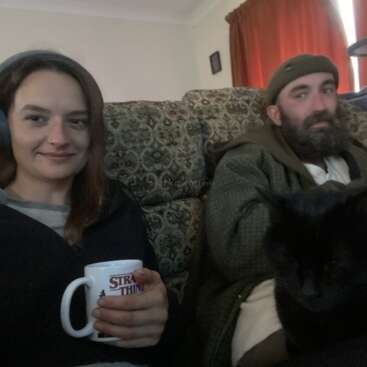 A woman wearing headphones and holding a "Stranger Things" mug sits on a patterned couch beside a bearded man and a black cat. Cozy atmosphere indoors.