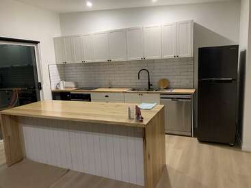 Esta imagen muestra una cocina moderna con armarios blancos, un grifo negro, electrodomésticos de acero inoxidable, una gran isla de madera y un frigorífico negro. Limpia y minimalista.