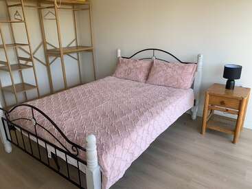 Esta imagen muestra una cama bien hecha con una colcha rosa y dos almohadas, una mesilla de noche de madera con una lámpara y una estantería abierta de madera.
