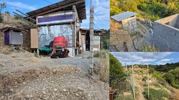 This image shows a rural, fenced farm area with sheds, a dog resting on the ground, various structures, and hilly, green surroundings under a blue sky.