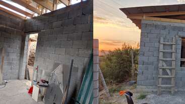 The image shows a small building under construction with grey concrete blocks, wooden roof framing, tools inside, and a wooden ladder outside at sunset in a rural setting.