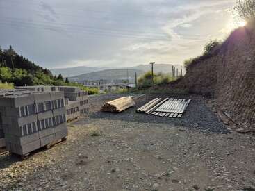 The image shows a construction site with stacks of concrete blocks, wooden planks, and metal bars, surrounded by hills and trees under a partly cloudy sky.