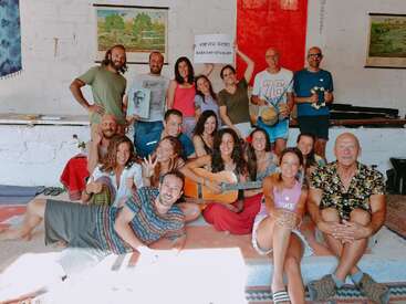 A joyful group of people gather indoors, some sitting, some standing. Several smile, one holds a guitar, another a tambourine. Relaxed, friendly atmosphere with colorful decor.