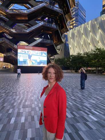 Eine Frau in einem roten Blazer steht selbstbewusst auf einem Platz in der Abenddämmerung. Hinter ihr sind moderne Gebäude, ein großer Bildschirm und das Gebäude The Vessel in New York City zu sehen.