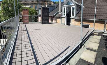 Dieses Bild zeigt eine geräumige Dachterrasse mit hellen Belägen, einem Metallgeländer, zwei Schornsteinen und einem kleinen Gebäudeeingang, umgeben von Nachbargebäuden und Grünflächen.