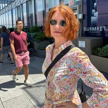 Eine Frau mit kurzen roten Haaren und Sonnenbrille posiert selbstbewusst im Außenbereich des Summit One Vanderbilt. Sie trägt ein bunt gemustertes Shirt, hinter ihr stehen Menschen und Pflanzen.