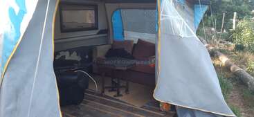 Esta imagen muestra el interior de una tienda o caravana improvisada. Se ven un sofá, un sillón, una mesita y una ventana, rodeados de vegetación exterior.