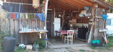 Esta imagen muestra una cocina rústica de madera al aire libre con techo abierto, mesa y sillas, estanterías, un fregadero, vajilla y diversos utensilios domésticos organizados alrededor.
