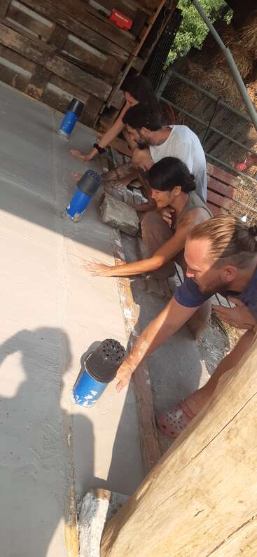Four people are smoothing out freshly poured concrete with their hands, working together on a floor inside a rustic wooden structure under the sunlight.