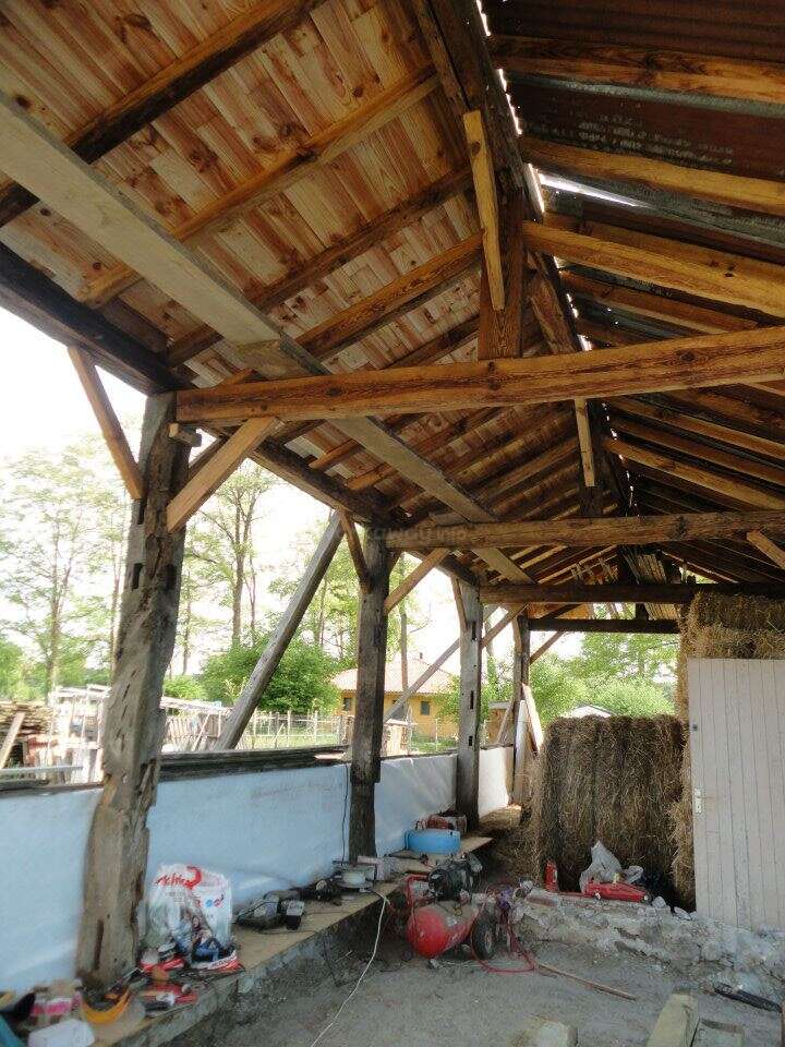 Esta imagen muestra el interior de un granero rústico con vigas de madera vistas, herramientas esparcidas, balas de heno, una pared parcialmente encalada y luz natural que se cuela por los huecos.