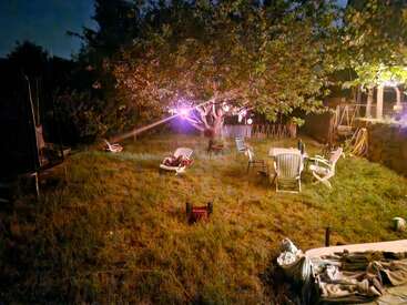 Eine nächtliche Szene im Garten, beleuchtet von sanften Lichtern. Gartenstühle, ein Baum, verstreute Gartenmöbel und ein Spielzeugmäher schaffen eine gemütliche, entspannte Atmosphäre auf der Wiese.