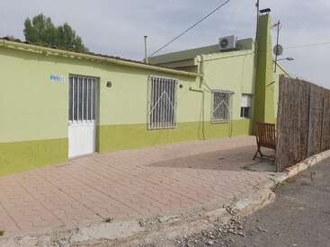 The image depicts a small, one-story building with a pale green exterior and a white door, featuring a paved walkway and a wooden bench outside.