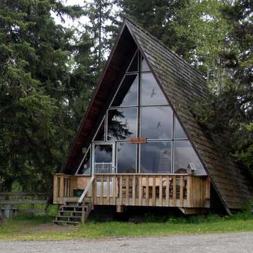 Esta imagen muestra una encantadora cabaña en forma de A con grandes ventanas frontales, terraza de madera, rodeada de altos árboles verdes y enclavada tranquilamente en una zona boscosa.