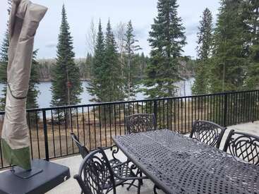 Une table et des chaises de jardin en métal sont disposées sur une terrasse en béton, avec un parasol recouvert d'une toile, donnant sur des pins, un lac et un ciel nuageux en arrière-plan.