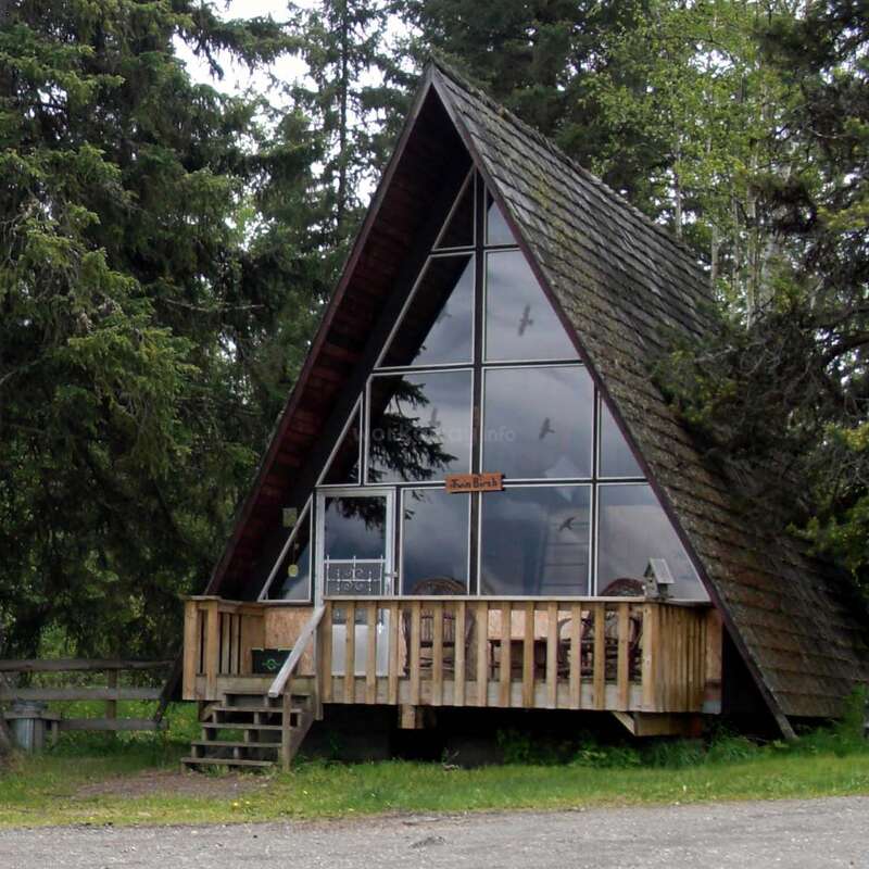 Esta imagen muestra una encantadora cabaña en forma de A con grandes ventanas frontales, terraza de madera, rodeada de altos árboles verdes y enclavada tranquilamente en una zona boscosa.