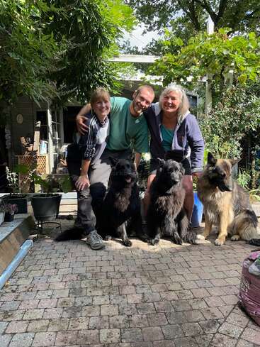 Trois personnes souriantes sont agenouillées à l'extérieur sur un patio en pierre, posant joyeusement avec trois grands chiens pelucheux. Une verdure luxuriante et une maison se trouvent à l'arrière-plan.