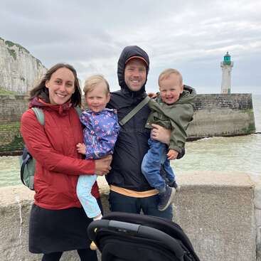 La imagen muestra a una familia de cuatro miembros posando para una foto en un muelle de hormigón, con un faro y un acantilado al fondo.