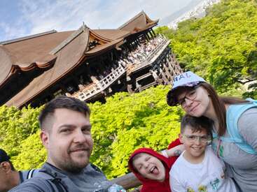 A family poses happily in front of a traditional Japanese temple surrounded by lush green trees. The children are smiling, making the scene joyful and lively.