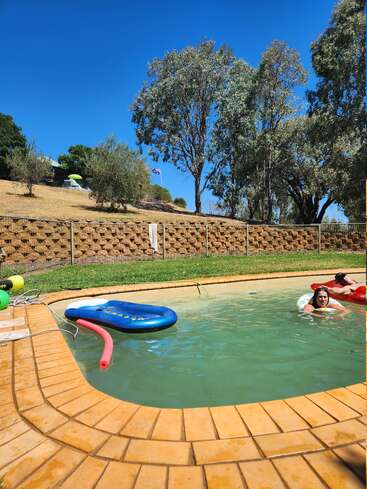 Um dia ensolarado na piscina com duas pessoas nadando, boias flutuando, cercadas por árvores e uma cerca, sob um céu azul claro. Atmosfera relaxante.