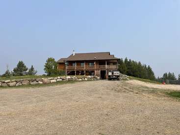The image depicts a two-story log cabin with a brown roof and balcony, situated on a dirt road, surrounded by trees and a stone wall, set against a clear blue sky.