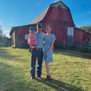 The image depicts a family of three standing in front of a rustic red barn on a sunny day, with the man holding a baby and the woman wearing a blue dress.