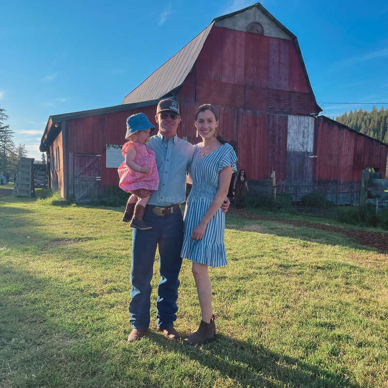 The image depicts a family of three standing in front of a rustic red barn on a sunny day, with the man holding a baby and the woman wearing a blue dress.