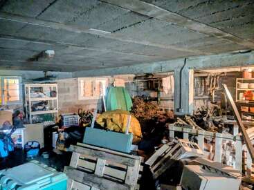 Dieses Bild zeigt einen überfüllten Keller oder eine Garage, vollgestopft mit Paletten, Säcken, Kisten, Werkzeugen, Schutt, Regalen mit allerlei Krimskrams und schwachem Tageslicht, das durch kleine Fenster hereinfällt.