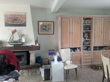 Ein gemütliches Wohnzimmer mit Holzschränken, Regalen und einem Kamin. Ein Modellsegelboot, gerahmte Bilder, Stühle, Haushaltsgegenstände und ein bisschen Unordnung sorgen für eine wohnliche Atmosphäre.