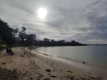 Das Bild zeigt eine friedliche, bewölkte Strandszene mit zwei Menschen, die am Ufer sitzen, mit aufgestellten Angelruten, ruhigem Wasser und Häusern, die sich in der Ferne als Silhouetten abzeichnen.