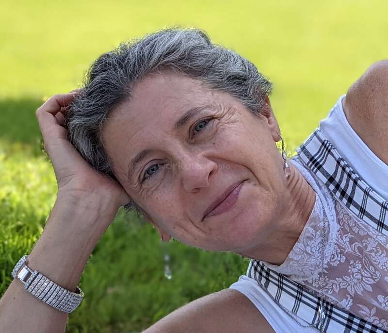 Das Bild zeigt eine ältere Frau mit kurzen grauen Haaren, die ein weißes Oberteil mit einem schwarz-weiß karierten Muster trägt und mit der Hand auf dem Kopf für die Kamera posiert.