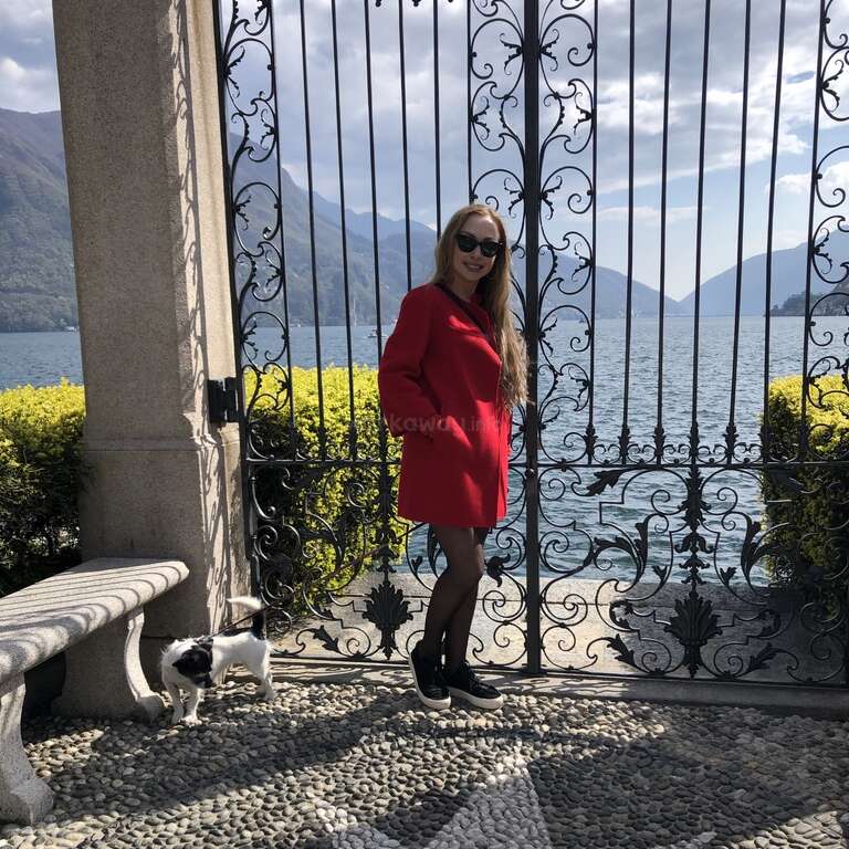 La imagen muestra a una mujer con un abrigo rojo delante de una verja ornamentada, con un perro y un banco cerca, en el marco de un lago y una montaña.