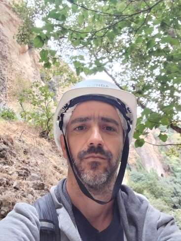 A man with a beard is wearing a white safety helmet and gray jacket, standing outdoors among trees and rocky cliffs, possibly hiking or climbing in nature.