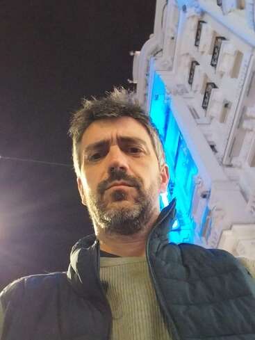 A man with dark hair and beard stands outdoors at night, wearing a jacket. Behind him, a decorative building glows with bright blue and white lights.