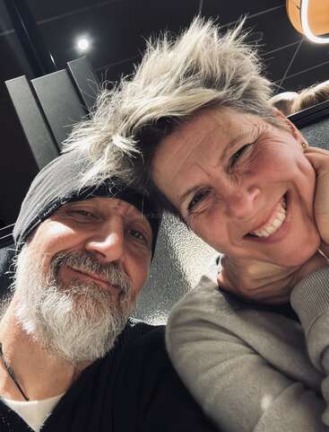 A smiling couple poses closely together. The man has a gray beard and wears a beanie, while the woman has short, spiked hair and cheerful expression.